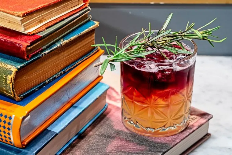 A glass of layered cocktail garnished with a rosemary sprig sits on a closed book, next to a stack of colorful, vintage books on a marble surface.