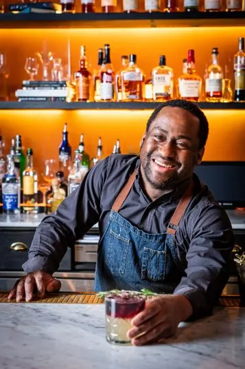 A smiling bartender in a dark shirt and denim apron stands behind a bar, offering a colorful cocktail. Shelves of liquor bottles are visible on a backlit orange wall.
