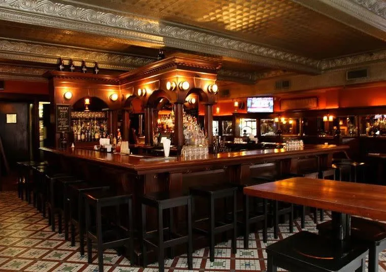 A warmly lit, empty bar with dark wood counters and black stools, ornate ceiling tiles, patterned tile floor, and shelves of liquor bottles behind the bar. A TV is visible in the background.