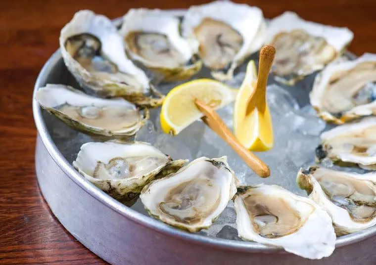 A metal tray of opened raw oysters on half shell arranged on ice, with two slices of lemon and a small wooden fork in the center, set on a wooden table.