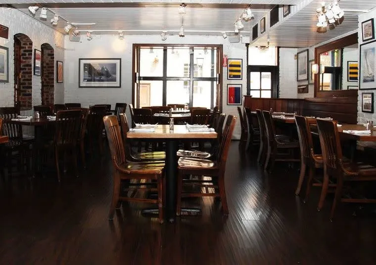A well-lit, empty restaurant dining area with dark wooden tables and chairs, white walls adorned with framed pictures, large windows, and wooden floors.