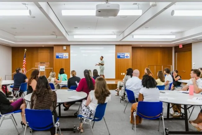 A speaker stands at the front of a conference room addressing an audience seated at round tables; United Way banners and an American flag are displayed on the stage behind the speaker.