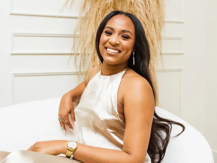 A woman in a shiny, sleeveless dress sits on a white chair, smiling. She has long, straight hair and wears a gold watch, earrings, and rings. Light decor and pampas grass are visible in the background.