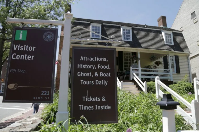 A brown visitor center sign and an attractions sign stand in front of a historic building with a porch, white railing, and people sitting outside on a sunny day.