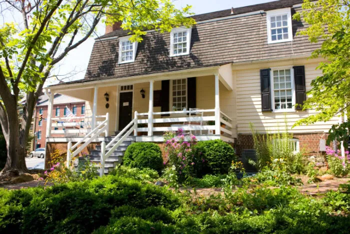 A historic yellow house with a dark brown gambrel roof, white railing, and shuttered windows, surrounded by lush landscaping and blooming flowers on a sunny day.