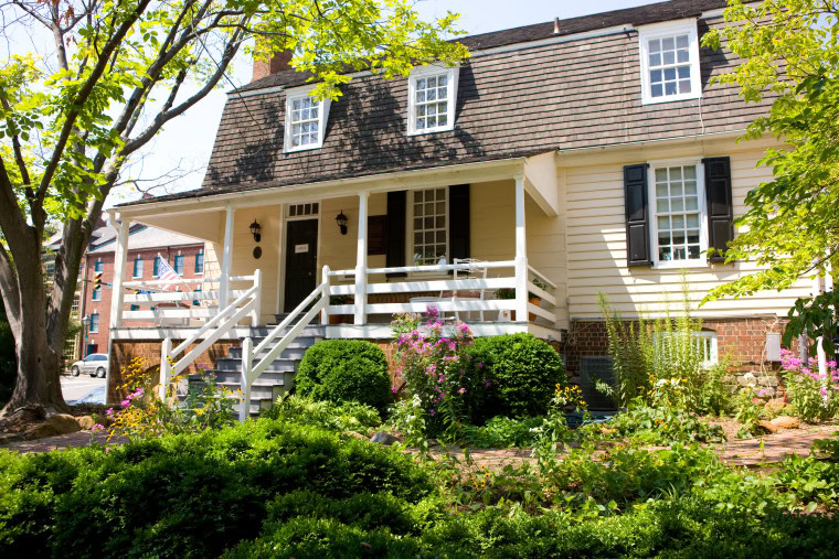 A historic yellow house with a dark brown gambrel roof, white railing, and shuttered windows, surrounded by lush landscaping and blooming flowers on a sunny day.