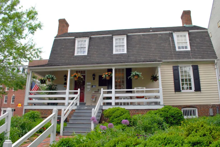 A two-story historic house with a dark gambrel roof, dormer windows, pale yellow siding, a front porch with hanging flower baskets, white railings, and a lush garden with green plants and purple flowers.