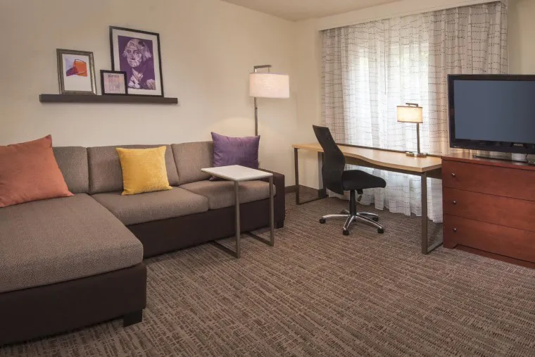 A modern hotel room with a sectional sofa, colorful pillows, framed art, a desk with a chair and lamp, a TV on a wooden dresser, and large window with sheer curtains letting in natural light.