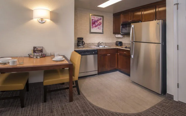A cozy kitchen and dining area with stainless steel appliances, wooden cabinets, a small table set for two, yellow chairs, and a coffee maker on the counter under a wall-mounted light.