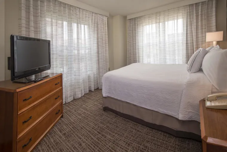 A hotel room with a large bed, two wooden dressers, a flat-screen TV, a bedside lamp, and floor-to-ceiling sheer curtains covering large windows. The room has neutral tones and carpeted flooring.