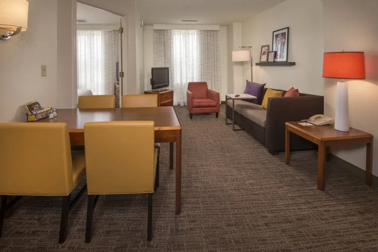 A hotel suite with a dining table and four yellow chairs, a brown sofa with colorful pillows, a red armchair, a TV, side tables with lamps, and large windows with sheer curtains.