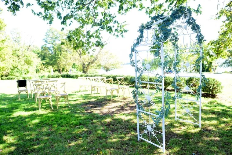 A white metal wedding arch decorated with green foliage stands on a grassy lawn under a tree, with several empty wooden chairs arranged in rows nearby. Sunlight filters through the leaves, creating a peaceful outdoor setting.
