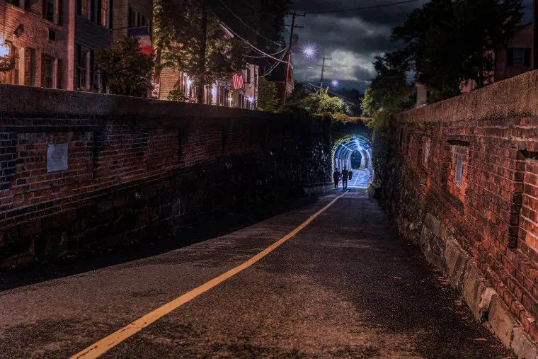 A dimly lit brick alleyway at night, with two silhouetted figures walking toward a glowing tunnel entrance. Streetlights and building windows illuminate the scene, casting long shadows on the wet pavement.