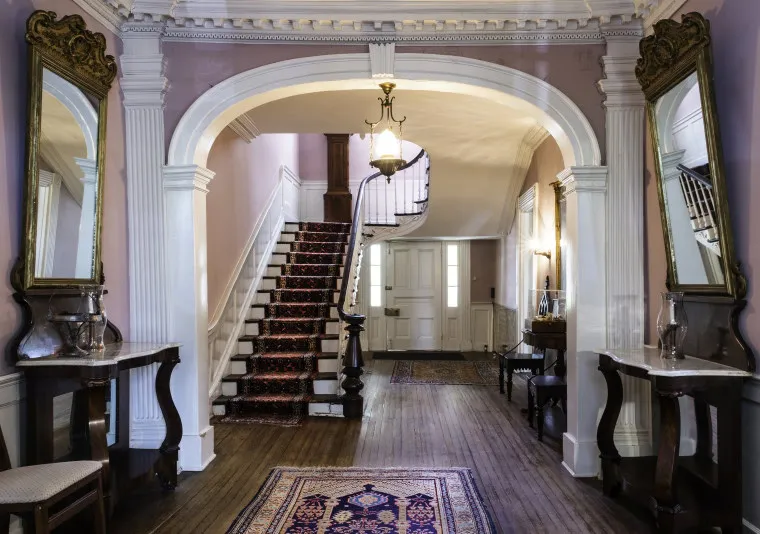 A grand hallway with wooden floors, ornate arches, two large mirrors, matching tables, and a red-carpeted staircase leading up. Walls are painted soft pink, with classic white trim and a chandelier overhead.