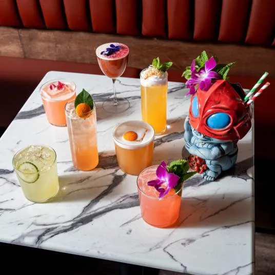 A marble table with seven colorful cocktails, each garnished with fruit or flowers, and a blue and red robot figurine holding straws, set against a red cushioned booth.