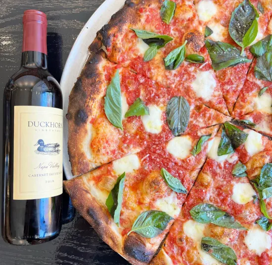 A bottle of Duckhorn Cabernet Sauvignon wine sits next to a margherita pizza topped with tomato sauce, melted mozzarella, and fresh basil on a dark table.