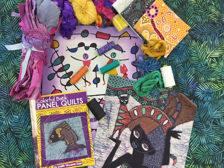 An assortment of colorful quilting materials, threads, batik fabrics, embroidery floss, a sewing pattern, and the book Colorful Batik Panel Quilts arranged on a leafy patterned fabric background.