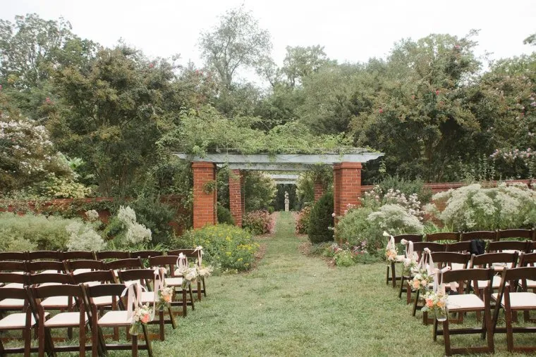 An outdoor wedding ceremony setup with rows of brown chairs decorated with flowers, facing a pergola covered in greenery and surrounded by lush gardens and trees.