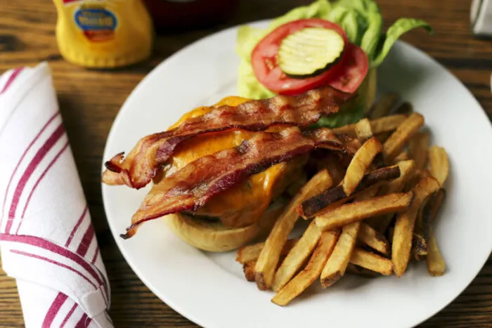 A plate with a bacon cheeseburger topped with bacon, cheddar cheese, lettuce, tomato, and pickles, served with a side of French fries. A wrapped napkin and a mustard bottle are nearby on a wooden table.