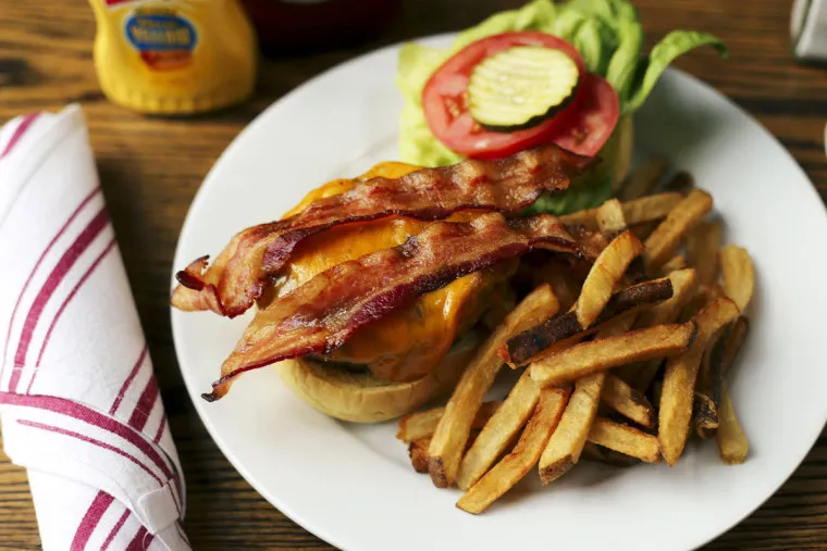 A plate with a bacon cheeseburger topped with bacon, cheddar cheese, lettuce, tomato, and pickles, served with a side of French fries. A wrapped napkin and a mustard bottle are nearby on a wooden table.