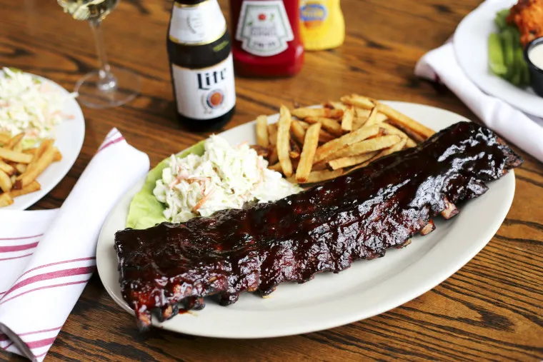 A plate of barbecued ribs with coleslaw and French fries on a wooden table, accompanied by a beer, condiments, and a side salad in the background.