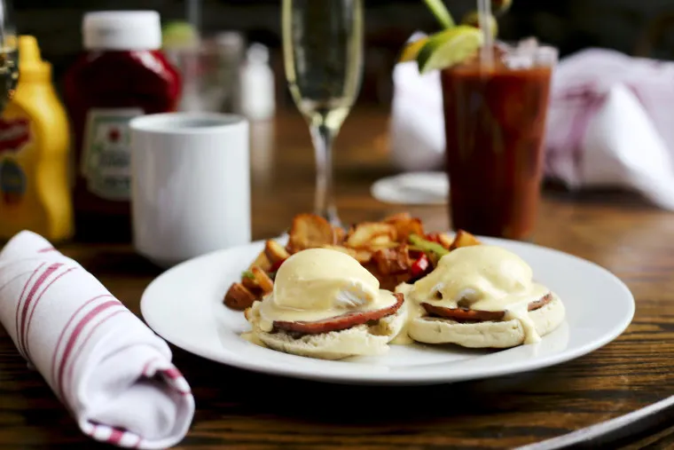 A plate of eggs Benedict with ham and hollandaise sauce sits next to breakfast potatoes. In the background are a cup, a Bloody Mary, a glass of champagne, and bottles of ketchup and mustard. A napkin is on the left.