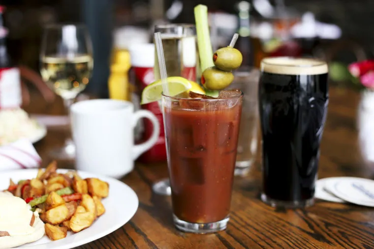 A Bloody Mary garnished with olives, a celery stick, and lime sits on a wooden table surrounded by a plate of food, a coffee mug, a glass of dark beer, a wine glass, and other drinks.