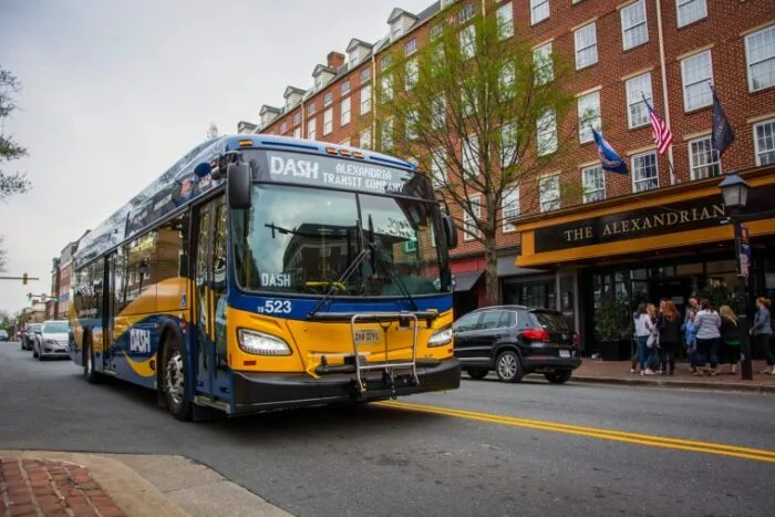 A DASH city bus drives past The Alexandrian hotel on a tree-lined street, with people gathered on the sidewalk and cars parked along the curb in Alexandria, Virginia.