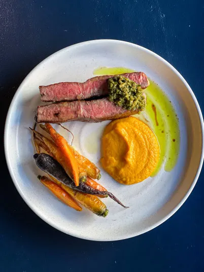 A plated meal with two slices of medium-rare steak topped with green sauce, a dollop of orange purée, and a small bunch of roasted multicolored carrots on a white plate against a dark blue background.