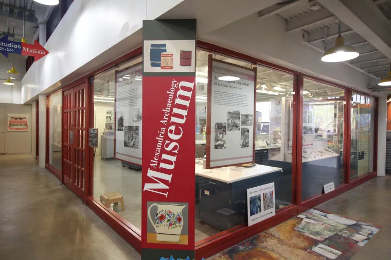 A glass-walled museum exhibit titled Alexandria Archaeology Museum with artifacts, informational displays, and photos inside; a red sign and a colorful map are visible in the foreground.