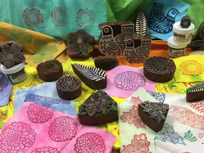 Wooden printing blocks with floral and bird designs, fabric paints, and colorful cloth pieces decorated with stamped patterns are arranged together on a vibrant background.