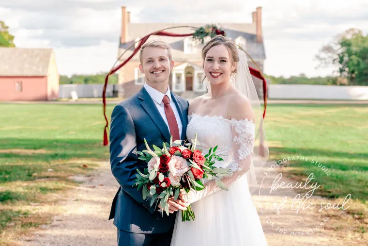 A bride and groom stand smiling outdoors in front of a farmhouse, holding a bouquet of red and white flowers. The bride wears an off-shoulder white dress and veil, and the groom wears a blue suit with a red tie.