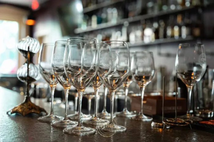 A row of empty wine glasses is neatly arranged on a bar counter, with shelves of liquor bottles and blurred background lighting creating a warm, inviting atmosphere.