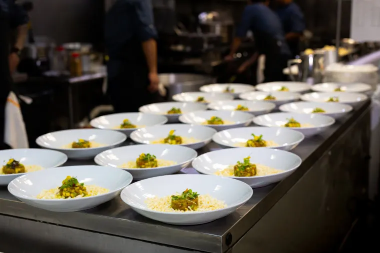 Several white bowls with plated food are arranged in rows on a stainless steel counter in a busy restaurant kitchen, with chefs working in the background.