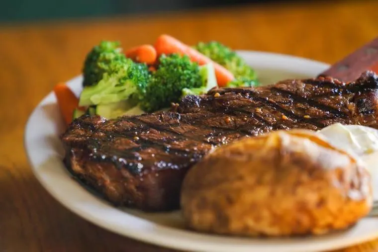 A plate of grilled steak with grill marks, served with a baked potato topped with sour cream, and a side of steamed broccoli and carrots.