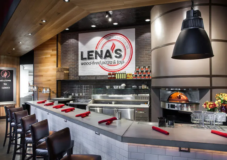 Modern pizza restaurant interior with a counter lined with chairs and red napkins, an open kitchen, a wood-fired pizza oven, and a large sign reading “Lena’s Wood-Fired Pizza & Tap” on the brick wall.