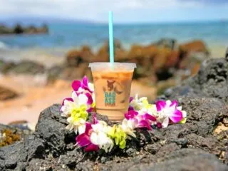 A plastic cup of iced coffee with a blue straw sits on black rocks, surrounded by a lei of purple and white flowers, with the ocean and sky blurred in the background.
