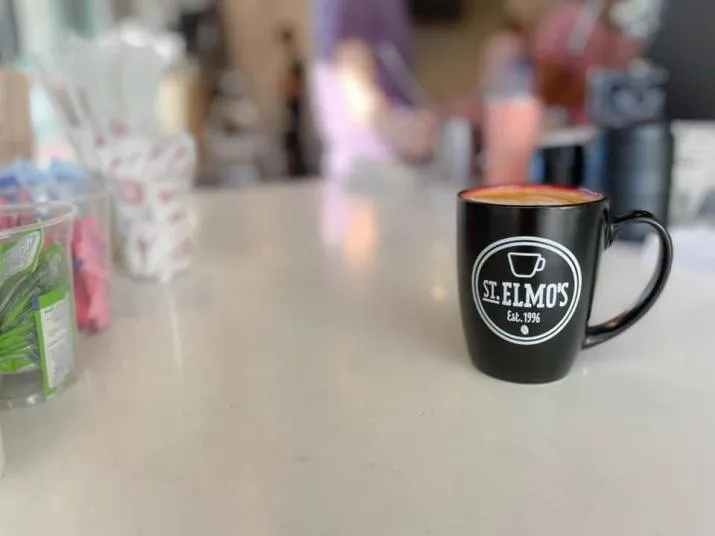 A black mug with St. Elmos Est. 1996 printed on it sits on a white countertop in a café, with blurred people and coffee shop items in the background.
