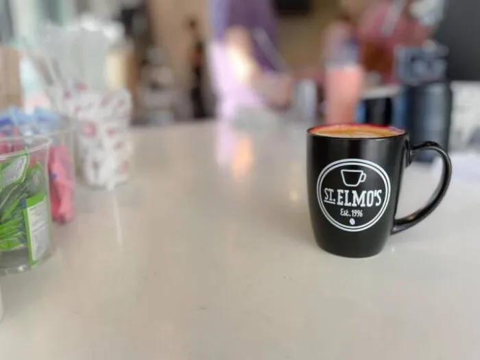 A black coffee mug with the St. Elmos logo sits on a white counter in a café, with blurred background figures and coffee shop items.