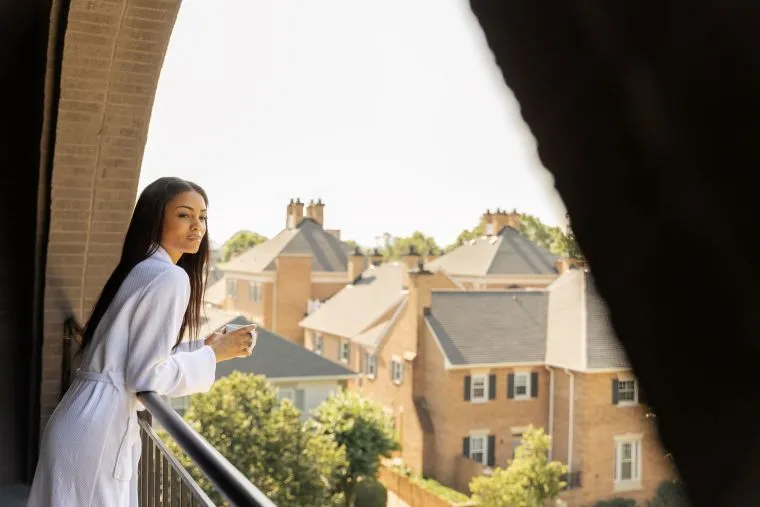 A woman in a white robe stands on a balcony, holding a mug and looking out over a neighborhood with brick houses and green trees on a sunny day.