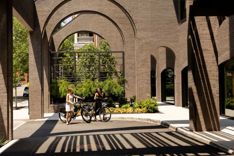 Two women stand with bicycles under large brick arches, talking in a sunlit, modern outdoor space with greenery and shadows from the arches falling across the ground.