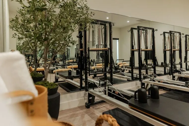 A modern Pilates studio with black reformer machines lined up in front of a large wall mirror. A potted plant and rolled white towels are visible in the foreground, creating a calm and inviting atmosphere.