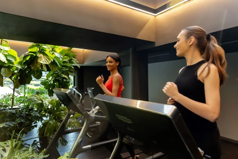 Two women exercising on treadmills in a modern gym with large green plants nearby, natural light coming in from the windows, and both women appearing focused and energetic.
