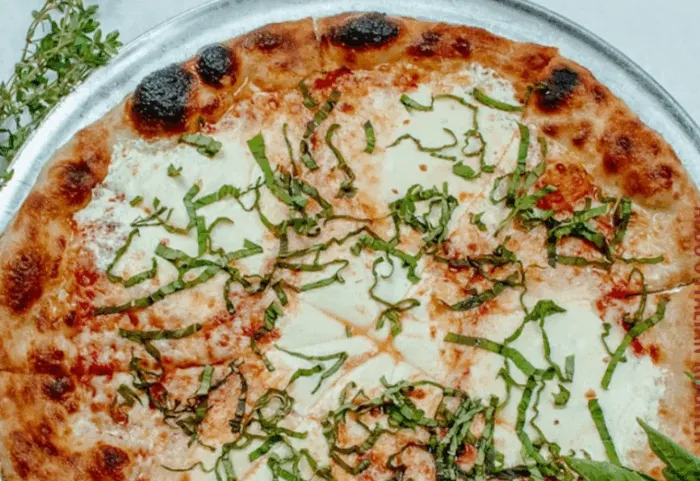 A close-up of a cooked pizza on a metal tray, topped with melted mozzarella cheese, tomato sauce, and freshly chopped basil leaves. The crust is golden-brown with some charred spots.