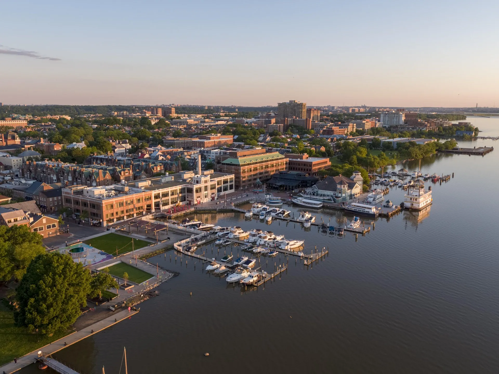 Aerial view of a marina with boats docked along the waterfront, surrounded by buildings, trees, and a park area at sunset in an urban setting.