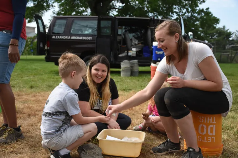 A woman kneels on a bucket, smiling and handing an object to a young boy at an outdoor event. Another woman sits nearby, smiling. A black van labeled Alexandria Archaeology is parked in the background.