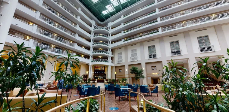 Spacious hotel atrium with several floors of balconies, indoor plants, blue and tan lounge chairs, small tables, and a bar area beneath a large glass ceiling letting in natural light.