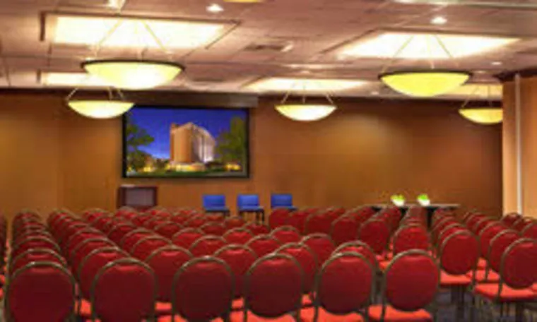 Rows of empty red chairs are arranged facing a stage with three blue chairs and a table, under round ceiling lights, in a conference room with a large screen displaying a building at night.