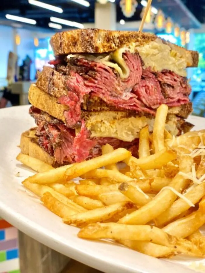 A close-up of a Reuben sandwich stacked with layers of corned beef, sauerkraut, and cheese on toasted rye bread, served with a generous portion of golden French fries on a white plate.