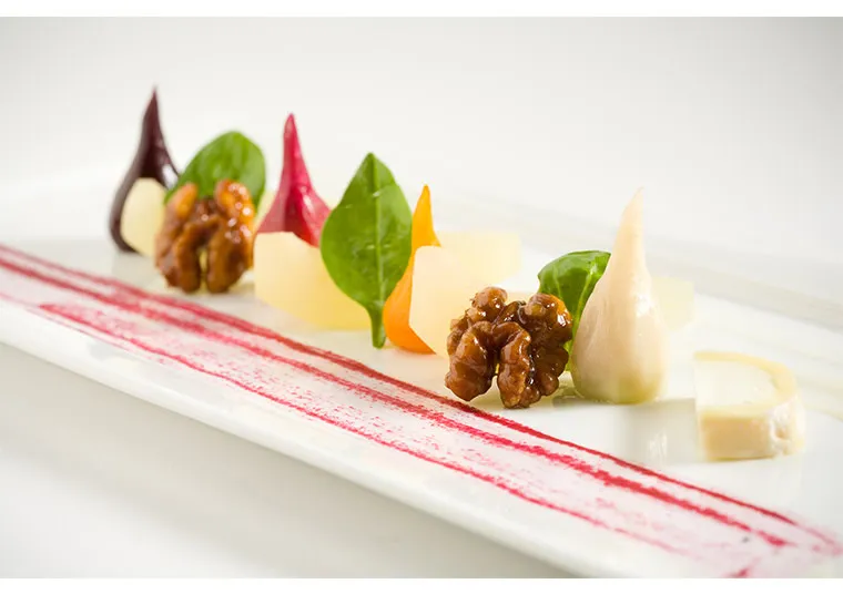A gourmet dish artfully arranged with colorful vegetables, walnuts, leafy greens, and cheese on a white plate, featuring a pink sauce streak for elegant presentation.
